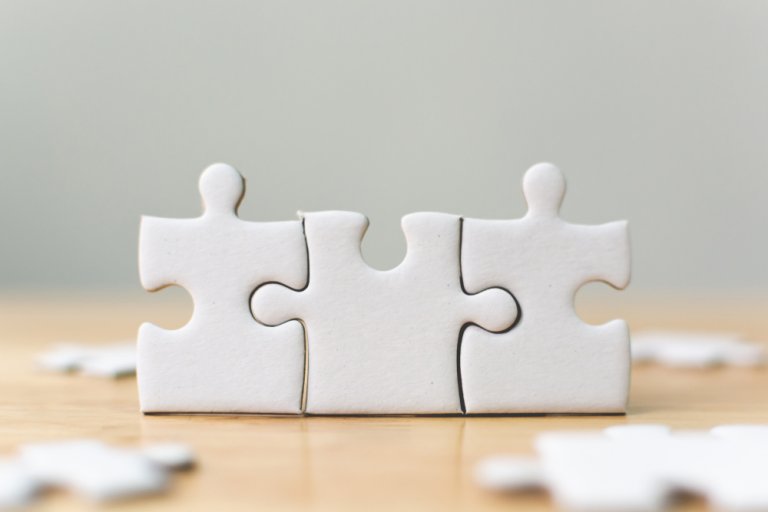 Three white jigsaw puzzle pieces are interlocked and standing upright on a light wood surface, with additional pieces scattered around in the background. The image evokes themes of problem-solving and connection, much like recruiters piecing together talent to form cohesive teams.