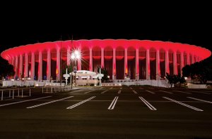 A large arena with distinctive white columns and a circular structure is illuminated by red lights against a dark night sky. The building brand is evident as the foreground features an empty parking lot with clearly marked lines and a few small trees on either side.