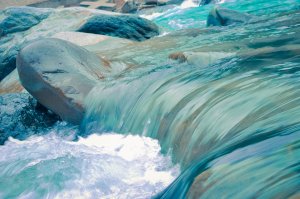 A serene stream of turquoise water flows over smooth, large rocks. Occasionally, whitewater ripples gently, creating a calming scene. The rocks, with their rounded shapes, add texture and contrast against the flowing water, enhancing the natural tranquility of the setting.