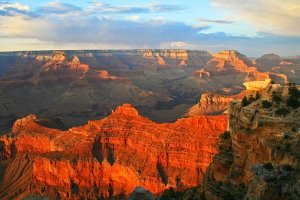 A stunning view of the Grand Canyon during sunset. The golden and orange hues reflect off the layered rock formations and deep valleys, contrasting against the partially cloudy blue sky. The landscape stretches out, showcasing the vastness and rugged beauty of the canyon, with whitewater rapids visible below.