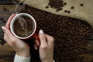 A person holding a red cup of steaming coffee with both hands. The cup is placed on a wooden surface covered with roasted coffee beans, evoking the rich culture behind every brew. A burlap sack, partially spilling coffee beans, is also visible.
