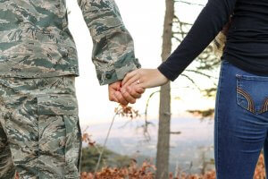 A person in a military uniform and another in casual attire hold hands outdoors, with trees and a blurred landscape in the background. Both individuals are seen from the back, focusing on their joined hands.