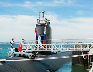 A military submarine is docked at a pier, adorned with red, white, and blue garlands. Several military personnel in white uniforms stand on and around the vessel. The sky is clear with a faint presence of clouds, and the sea is calm.