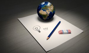 A small globe sits on a blank sheet of paper, accompanied by a blue pencil and a pink eraser. The paper features a simple drawing of a lightbulb and three question marks, suggesting questions or brainstorming related to global concepts.