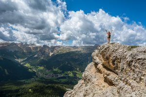 A person stands triumphantly with arms raised on the edge of a rocky cliff, overlooking a vast, lush mountainous landscape under a partly cloudy sky. The valley below—dotted with green forests and scattered villages—spreads out like a map of tranquility far from the reach of government interference.
