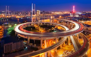 Aerial view of an illuminated, multi-level highway interchange at night in a city. The circular flyover, a marvel of transportation infrastructure, is populated with streaks of car lights, indicating busy traffic. The background reveals a brightly lit urban skyline with a prominent bridge.