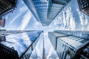 Low-angle shot of several modern skyscrapers with reflective glass surfaces converging towards the sky. The buildings stretch upwards surrounded by a blue sky with scattered, wispy clouds. The reflections on the glass showcase parts of the adjacent buildings and clouds above.