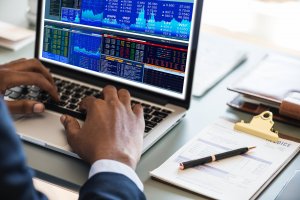 A person is using a laptop displaying multiple financial charts and data tables. Nearby, there is a notepad with a pen, a clipboard with documents, and a pair of glasses on the desk. The scene appears to be a financial analysis or stock trading workspace leveraging cloud-based tools for real-time data.