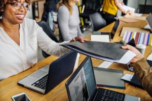 A smiling person seated at a table in an office hands a folder to another individual. The table, cluttered with laptops, paper, and various office supplies, showcases the hustle of the workspace. In the background, people are engaged in their work while discussing cloud solutions.