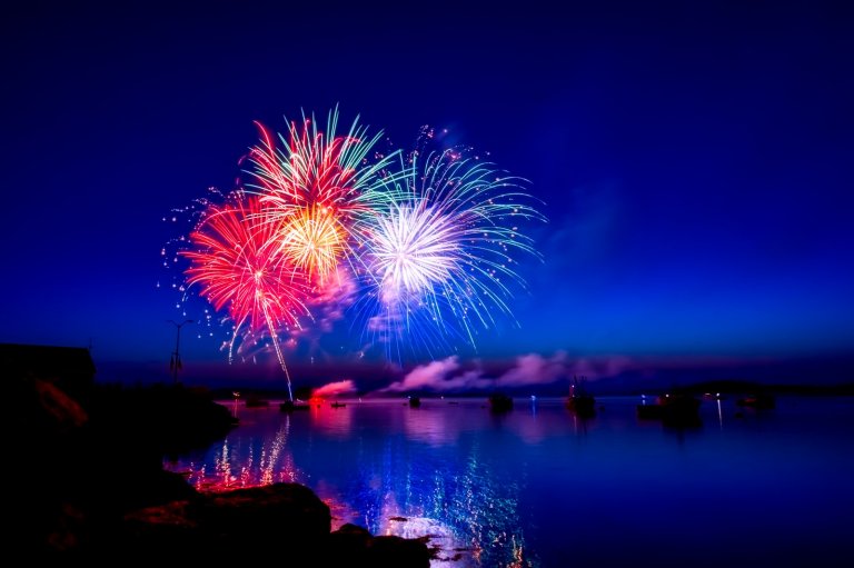 Colorful fireworks explode in the night sky, their vibrant red, green, and white bursts reflected on the calm water below. In the distance, government boats float on the water, and the horizon fades into a deep blue gradient.