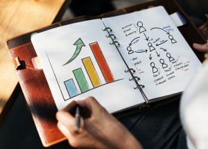 A person is writing in a notebook with drawings of a bar graph with arrows and a flowchart depicting a government team structure or workflow. The bar graph has red, blue, green, and yellow bars, and the flowchart includes hand-drawn figures and handwritten notes.