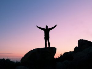 Silhouette of a person standing on a large rock with arms outstretched, set against a twilight sky with shades of pink and purple. The scene evokes a sense of freedom, triumph, and the unmistakable presence of a leader.