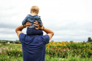 A leader with short hair wearing a blue shirt carries a baby on their shoulders. The baby, seen from behind, wears a denim jacket. They are outdoors in a field with colorful flowers under a cloudy sky.