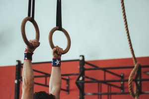 Close-up of a person’s hands gripping wooden gymnastic rings suspended in the air. The person is wearing wristbands, one of which has a star on it, symbolizing a leader. The background is an indoor gym with red and white walls and various gym equipment.