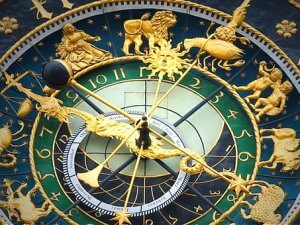 An intricate astrological clock with gold and emerald green details. The clock, known for its resistance to the elements, features zodiac symbols, an ornate sun in the center, and various celestial designs. The hands of the clock are elaborately decorated, and the background shows Roman numerals.