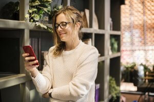 A woman wearing glasses and a white sweater is smiling while looking at her phone, likely engaging in some networking. She is standing in front of a bookshelf with plants and various items. The background features a mix of natural light and shadows.