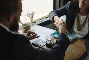 A man and a woman are seated at a round wooden table, shaking hands in a professional gesture of networking. They are both wearing business attire. On the table, there is a cup of black coffee, a green saucer, a folder with papers, and a small decorative plant in a jar.