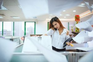 A woman in a white blouse stands in a busy office, deftly navigating the copier with impressive soft skills. Papers are flying around the room, some slightly blurred due to motion. The office has cubicles and large windows in the background, allowing natural light to enter.