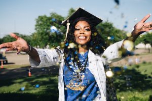 A joyful person in a graduation cap and gown throws blue and gold confetti in the air. They are outdoors on a sunny day with trees and grass in the background, smiling broadly and looking directly at the camera, showcasing the soft skills they've honed throughout their academic journey.