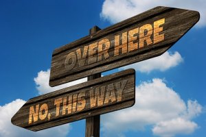 Two wooden directional signs against a blue sky with clouds. The top sign points left and reads "OVER HERE," while the bottom sign points right and reads "NO, THIS WAY." It's like a perfect metaphor for over-thinking—going back and forth without ever making a decision.