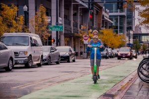 A young boy and an older boy ride together on an electric scooter in a city bike lane, enjoying their mode of transportation. Autumn trees line the street, with cars and buildings visible in the background. The street sign reads "Idaho.