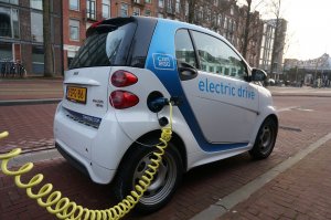 A white and blue electric car, a modern marvel of transportation, is parked on a brick road, plugged into a yellow charging cable connected to a charging station. The side of the car proudly displays the text "electric drive." Buildings and trees provide a serene background.
