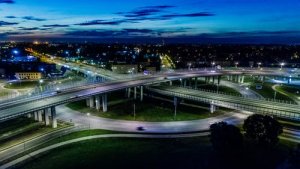 An aerial view of a brightly lit highway interchange at dusk showcases the intricate pattern of overpasses and underpasses central to modern transportation. Surrounding city lights and buildings add to the scene as the sky transitions to night.