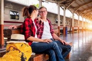 A young couple sits on a bench at a train station, casually dressed in red flannel shirts. The woman’s yellow backpack and straw hat rest beside her as the man wraps his arm around her. A train is visible in the background while sunlight streams in, creating a scene that speaks to employee engagement and togetherness.