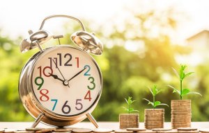 An analog alarm clock with colorful numbers sits on a wooden surface. Beside it, three ascending stacks of coins, each topped with a small growing plant, symbolize financial growth and employee engagement over time. The background is blurred, featuring greenery and sunlight.