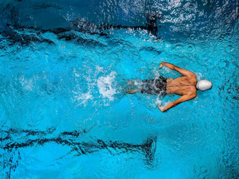 A swimmer wearing a white swim cap and goggles performs a stroke in clear blue water, reminiscent of military precision. The image taken from above captures the swimmer mid-swim, creating splashes as shadows of lane dividers line the pool floor.