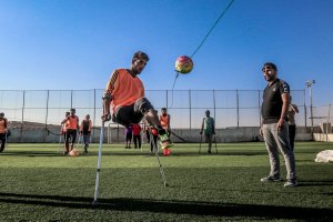 A soccer player with a prosthetic leg skillfully kicks a ball while others, some also with military backgrounds and prosthetic limbs, watch and wait on a grass field. The background shows a clear blue sky and a fence enclosing the field. One coach or referee stands nearby observing.
