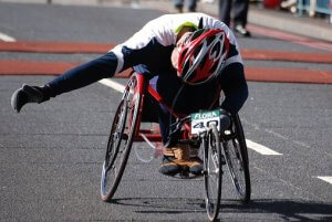 A person wearing a helmet and racing gear is competing in a wheelchair race. They are leaning forward intensely with one arm outstretched and the other on the wheel, showing military-like determination and athleticism on a city street.
