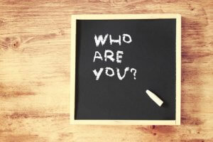 A small blackboard on a wooden surface with the words "WHO ARE YOU?" written in white chalk. A piece of white chalk lies on the bottom right corner of the blackboard, inviting you to define your brand.