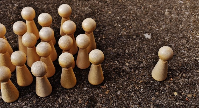A group of light wooden pawns stands densely together on a textured dark surface while a single pawn is isolated from the group at a short distance away, evoking themes of individuality and separation, reminiscent of bullying.