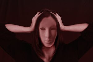 A person with a featureless mask holds their head with both hands, set against a dark, red-tinged background, evoking the torment often experienced by victims of bullying. The scene conveys an intense and emotional atmosphere.