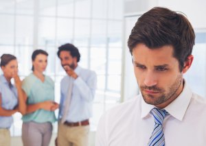 A man in a white shirt and blue striped tie looks downcast in the foreground. In the background, three colleagues—two women and one man—stand together, conversing and smiling. The scene subtly hints at bullying, with their eyes seemingly on the isolated man.
