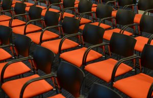 Rows of empty chairs with black backs and armrests and bright orange seats, arranged in a grid pattern on a dark carpeted floor, create a visually striking and orderly arrangement, perfect for an industry event.