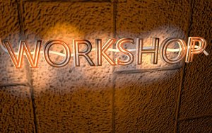 The image shows the word "WORKSHOP" in bright, neon-lit letters against a textured, brown-orange surface, possibly a wall. The neon glow creates a warm, illuminated effect around the letters, evoking the vibrant ambiance of an industry event.