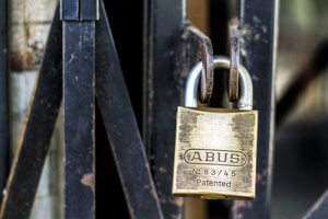 Close-up image of a brass padlock labeled "ABUS Patented N-83/45" securing a metal gate. The gate is made of rusted metal bars overlapping at various angles, contributing to an industrial and weathered appearance, symbolizing strong cybersecurity measures. The focus is sharp on the padlock and surrounding gate area.