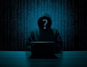 A person wearing a hooded sweatshirt sits in front of a laptop in a dark room, with a large question mark covering their face. The background features rows of digital code, giving the image a mysterious and cyber-themed atmosphere, reflecting the elusive world of cybersecurity.