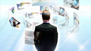 A man in a suit stands facing a curved array of floating screens and holographic images depicting technology, digital content, and cybersecurity, set against a bright, futuristic background. The display includes icons, graphs, and globe imagery, suggesting a focus on global data and innovation.