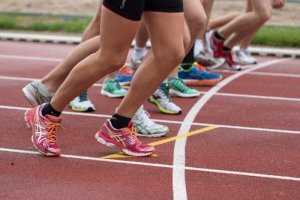 Several runners are poised at the starting line of a race track, ready to start a challenging downhill sprint. The focus is on their legs and running shoes, which are in various colors and designs, as they crouch down in anticipation of the race.