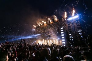 A large crowd attends a nighttime concert with a brightly lit stage. Flames and streamers shoot from the top of the stage, and attendees raise their hands, enjoying the energetic atmosphere. Set downhill, the dramatic backdrop of the dark sky only amplifies the event's intensity.