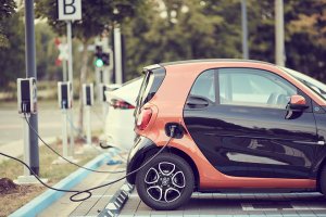 A small, two-toned electric vehicle is plugged into a charging station in a parking lot. Other electric cars are charging nearby. The background features blurred trees and a sign with the letter "B.