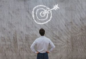 A person in a white shirt and jeans stands with hands on hips, facing a gray concrete wall with a white chalk drawing of a target and arrow hitting the bullseye, symbolizing achieving goals or meeting high expectations.