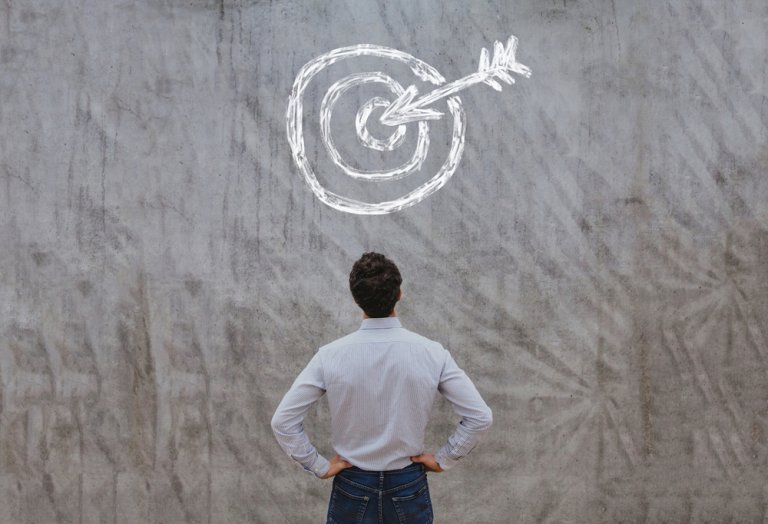 A person in a white shirt and jeans stands with hands on hips, facing a gray concrete wall with a white chalk drawing of a target and arrow hitting the bullseye, symbolizing achieving goals or meeting high expectations.
