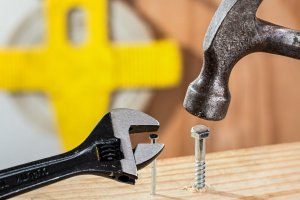 A wrench from a popular brand is mistakenly tightening a nail while a hammer is about to hit a bolt on a wooden surface. In the background, a yellow and gray blurred object is partially visible.