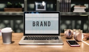 A laptop showcasing the word "BRAND" sits on a wooden table alongside a cup of coffee, a pair of headphones, and a smartphone. The cozy workspace is completed with shelves filled with books and a framed picture in the background.