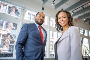 Two people are standing indoors, professionally dressed. The man on the left wears a blue suit and red tie, and the woman on the right wears a light gray blazer. Both are smiling proudly, as if celebrating a recent raise, with large windows showcasing the exterior of a building in the background.