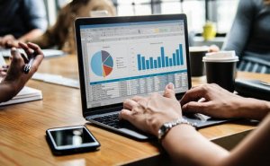 A person is using a laptop displaying a spreadsheet with colorful graphs and charts, including a pie chart and bar graph highlighting employee engagement metrics. A coffee cup, notebook, pen, and smartphone are on the wooden table. Other people are blurred in the background.